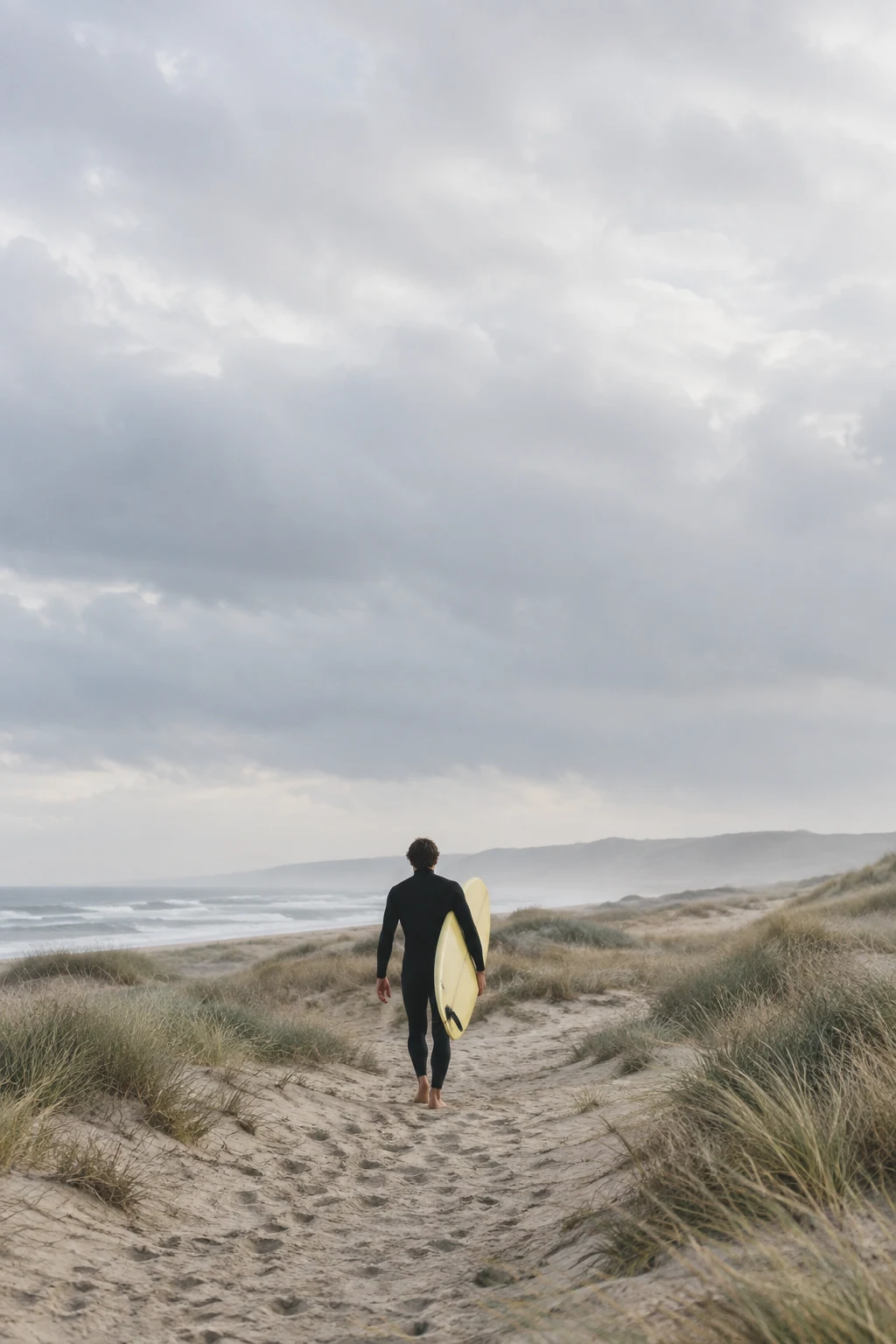 A lone surfer carries a pale board towards quiet dunes under a wide northern sky.