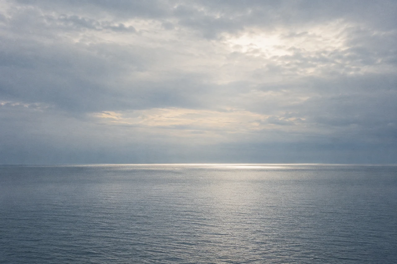 A single band of light crosses a quiet blue-grey sea beneath an expansive clouded sky.